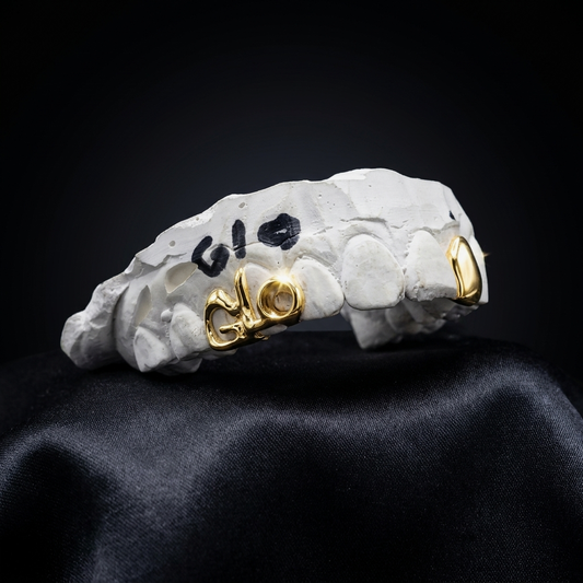 14K yellow gold "GLO" script lettering grillz paired with a solid incisor tooth cap on a plaster dental model over black satin.