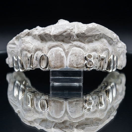Sterling silver double canine caps and custom cut-out number "1087" incisor grillz displayed on a plaster tooth model.