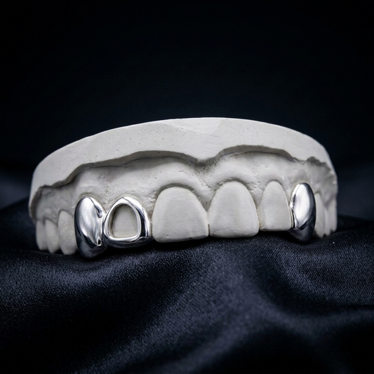 Custom silver/white gold 2-piece grillz set combining a solid full-coverage tooth cap with an open cutout window bar and a single canine cap. Precision custom dental jewelry.Silver open-window bar and solid tooth cap 2-piece grillz custom fitted on a plaster dental mold over dark fabric.